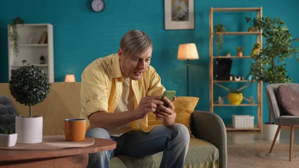 一个男人坐在沙发上 用智能手机关了起来 一个男人通过社交网络浏览他的信息 看照片 玩网络游戏 家庭幸福的概念 — 图库照片