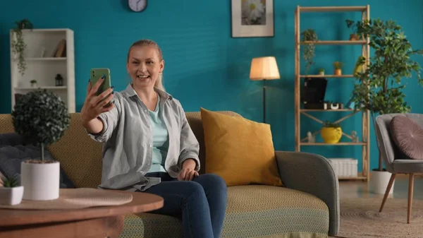 女人在智能手机上拍自拍或通过视频通话 沙发上客厅里拿着电话的女人的画像 — 图库照片