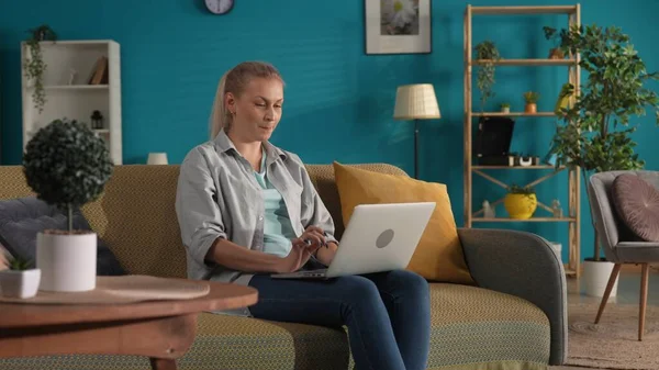 カジュアルな服を着た女性がリビングルームのソファーに座り ラップにラップトップを貼っています 女性は働き テキスト メッセージ チャット オンラインゲームをタイプします リモートワーク フリーランサー — ストック写真