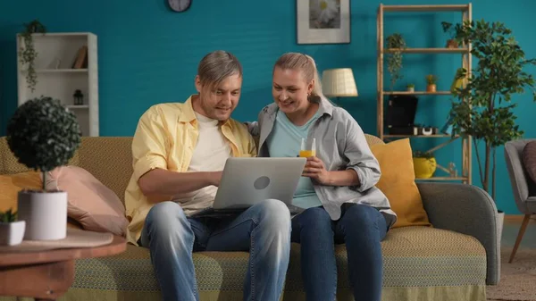 A man and a woman are watching a movie or video on a laptop. Portrait of a married couple in the living room on the sofa, the man has a laptop on his lap, and the woman is holding a glass of juice