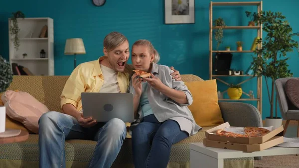 A man and a woman watch a movie, video on a laptop and eating pizza. Portrait of a family sitting in an embrace on the sofa in the living room with a laptop on her lap. Family idyll