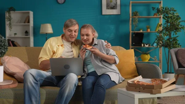 一个男人和一个女人在看电影 在笔记本电脑上的视频和吃披萨 在客厅的沙发上 一家人抱着她坐在沙发上 膝上型电脑的肖像 家庭田园诗 — 图库照片