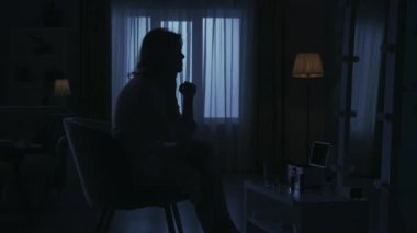 Karanlık dairedeki kadın siluetinin portresi. Günlük yaratıcı yaşam konsepti. Bornozlu kadın odada bir sandalyede oturuyor, aynaya bakıyor, telefonda biriyle konuşuyor..