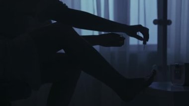 Karanlık dairedeki kadın siluetinin portresi. Günlük yaratıcı yaşam konsepti. Bornozlu kadın odada bir sandalyede oturuyor, tırnak cilası alıyor, bacağı masanın üstünde, pedikür yapmaya başlıyor..