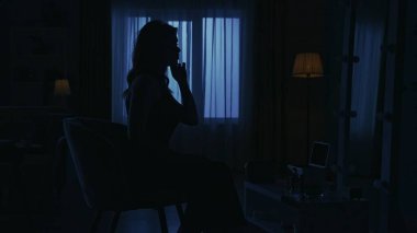 Karanlık dairedeki kadın siluetinin portresi. Günlük yaratıcı yaşam konsepti. Elbiseli ve topuklu ayakkabılı bir kadın odada bir sandalyede oturuyor, aynada kendine bakıyor, yüzüne dokunuyor..