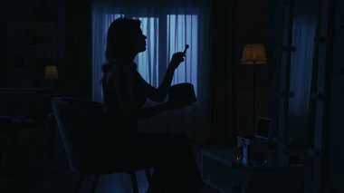 Karanlık dairedeki kadın siluetinin portresi. Günlük yaratıcı yaşam konsepti. Yakın çekim çekiyoruz. Odada bir sandalyede oturan, aynaya bakan, fırçayla makyaj yapan bir kadın..