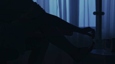 Karanlık dairedeki kadın siluetinin portresi. Günlük yaratıcı yaşam konsepti. Yakın plan çekim. Bornozlu kadın karanlık odada bir sandalyede oturuyor, bacağını kaldırıyor, pedikür yapıyor..