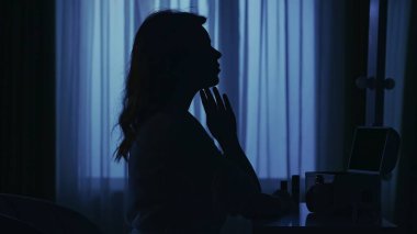 Karanlık dairedeki kadın siluetinin portresi. Günlük yaratıcı yaşam konsepti. Bornozlu kadın odada bir sandalyede oturuyor, aynaya bakıyor, yüzüne dokunuyor..