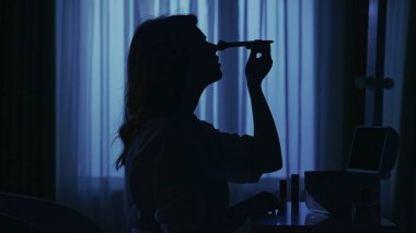 Karanlık dairedeki kadın siluetinin portresi. Günlük yaratıcı yaşam konsepti. Bornozlu kadın odada bir sandalyede oturuyor, aynaya bakıyor, fırçayla toz uyguluyor..