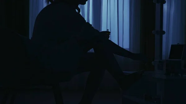 Portrait of female silhouette in the dark apartment. Everyday life creative concept. Woman in bathrobe sitting on a chair in the room legs up on the table, doing pedicure with nail polish.