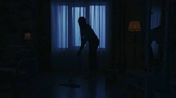 Portrait of female silhouette in the dark apartment room. Everyday life creative concept. Woman in casual clothing mopping the floor in the living room.