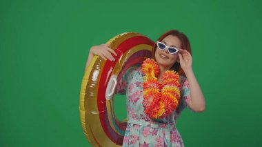 Gezici reklam yaratıcı konsepti. Gözlüklü ve çiçekli kadın gezgin elinde şişme tüp yüzük, tatilde eğleniyor, kameraya poz veriyor. Yeşil arkaplanda izole.