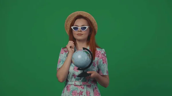 旅行広告クリエイティブコンセプト 小さな地球を保持するストロー帽子とメガネで女性旅行者 そして手を挙げてカメラにポーズ 緑の背景に孤立した — ストック写真