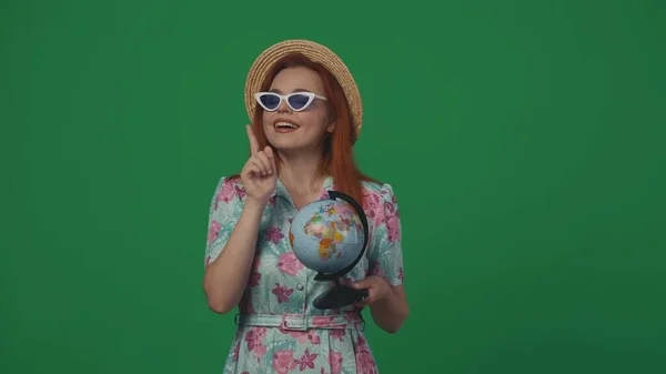 旅行广告创意的概念 戴着草帽和眼镜的女旅行者举着一个摆设在镜头前微笑着的面容的地球仪 被绿色背景隔离 — 图库照片