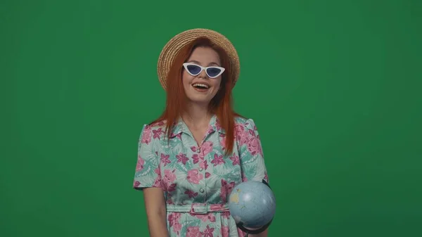 旅行广告创意的概念 戴着草帽和眼镜的女旅行者举着地球仪 看着摄像机快乐的面容 被绿色背景隔离 — 图库照片