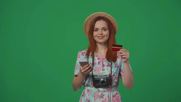 旅行广告创意的概念 头戴草帽的女旅行者手持信用卡和智能手机 在线支付服务 面带微笑 被绿色背景隔离 — 图库照片