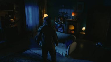 Korku filmi sahnesi. Cadılar Bayramı reklam konsepti. Elinde beyzbol sopası olan genç bir kız ve karanlık oturma odasında elinde bıçakla kanepede yürüyen beyaz maskeli bir adam..