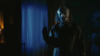 Korku filmi sahnesi. Cadılar Bayramı reklam konsepti. Beyaz hokey maskeli adam karanlık oturma odasında elinde bıçakla kameraya bakıyor..