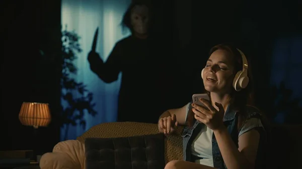 Horror movie scene. Halloween advertisement concept. Girl in headphones sitting on the sofa in the living room, listening to music, man maniac in white mask standing with knife near window behind.