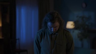 Daire yaratıcı konseptinde korku filmi sahnesi. Uzun saçlı adam karanlık oturma odasında dikiliyor, kameraya bakıyor, kendinden emin bir yüzle poz veriyor, dışarıda fırtına var. HDR