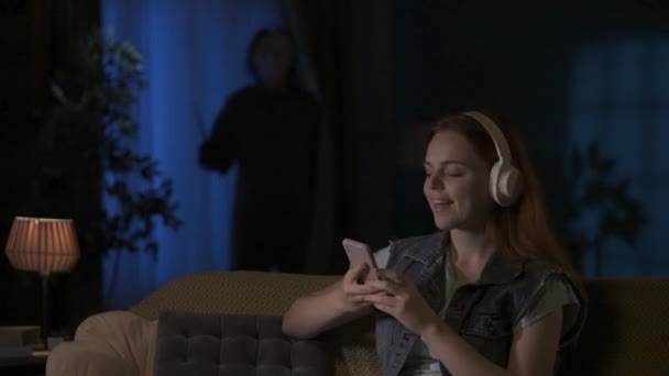 アパートのクリエイティブコンセプトのホラー映画シーン ダークリビングルームのソファーに座っているヘッドフォンの少女 音楽を聴くこと ナイフで隠れているマニアック 彼女の後ろに立っている — ストック動画
