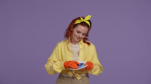 クリーニングとハウスキーピングのクリエイティブコンセプト カジュアルな服とゴム手袋の女性は プレートを保持し スポンジでそれを洗い カメラを見ている笑顔 紫色の背景に隔離される — ストック動画