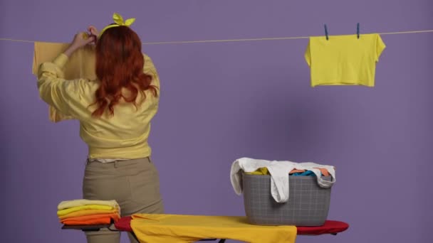 清洁和家政创意的概念 穿着休闲装的女人站在熨烫板旁边 篮子里装满了衣服 把衣服挂在绳子上 被紫色背景隔离 — 图库视频影像