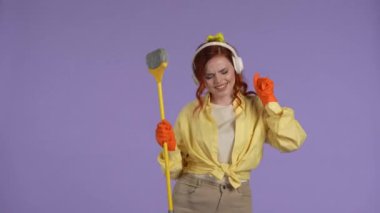 Temizlik ve temizlik yaratıcı konsepti. Günlük kıyafetler giymiş, kulaklıklı, süpürgeyle dans eden, müzik dinleyen, şarkı söyleyen, gülümseyen bir kadın. Mor arkaplanda izole.