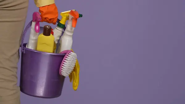 Everyday cleaning and housekeeping concept. Closeup shot of woman in casual clothing and rubber gloves holding bucket of cleansers, rags and brushes. Isolated on purple background.