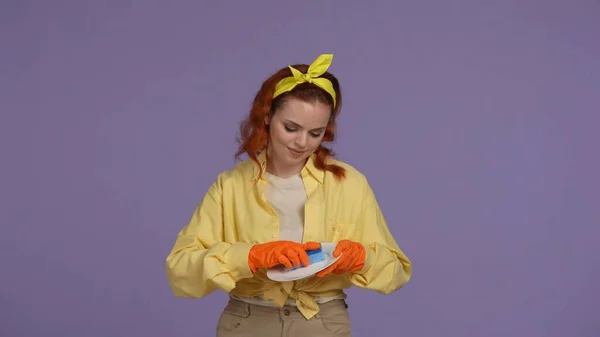 日常清洁和家务事的概念 穿着休闲装和橡胶手套的女人拿着盘子 用海绵擦拭 被紫色背景隔离 — 图库照片