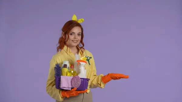 日常清洁和家务事的概念 穿着休闲装和橡胶手套的女人拿着装有清洁剂的篮子 用手显示空旷的区域 被紫色背景隔离 — 图库照片