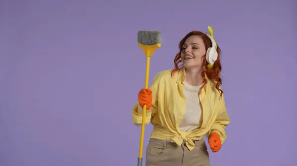 日常清洁和家务事的概念 穿着休闲装 头戴橡胶手套 手持扫帚 听音乐 面带微笑的女人 被紫色背景隔离 — 图库照片