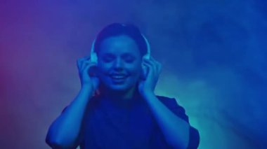 Beyaz kablosuz kulaklık takan genç, kızıl saçlı bir kadın DJ müziği dinliyor ve enerjik bir şekilde dans ediyor. Stüdyoda dans eden bir kadın renkli neon ışıklarıyla duman bulutları arasında