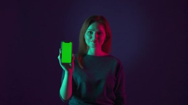 Bir kadın işaret parmağını akıllı telefonunun yeşil ekranına doğrultuyor ve baş parmağını doğrultuyor. Stüdyoda pembe arka planda yeşil ekranlı akıllı telefonlu bir kadının portresi.