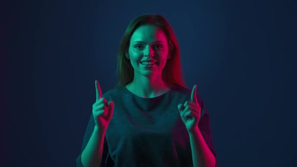 自信のある女性が指を指差して親指を示す ピンクと緑のネオンライトの青い背景にあるスタジオで赤毛の女性 — ストック動画