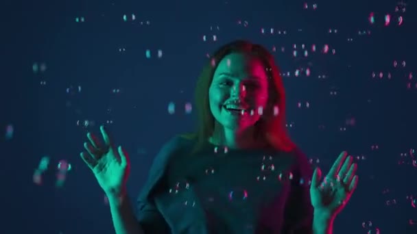快乐的女人喜欢用肥皂泡泡泡 泡泡泡和拍手 一个无忧无虑的女人的画像在工作室里被肥皂泡围绕着 蓝色背景上有粉色和绿色的霓虹灯 — 图库视频影像