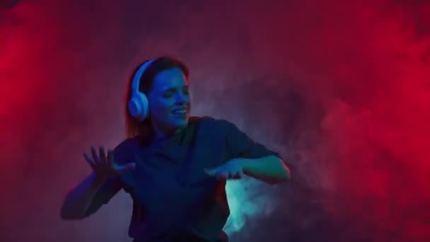 白いワイヤレスヘッドフォンを着用した若い赤毛の女性がDjの音楽を聴き 精力的に踊ります 女性は多色ネオンライトの煙の雲の中でスタジオで踊る — ストック動画