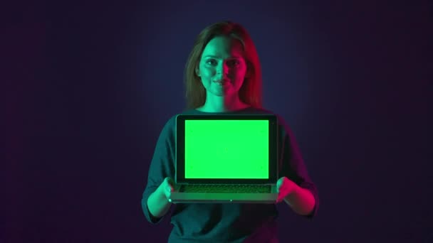 ノートパソコンの緑色の画面を閉じている女性 ピンクと緑のネオンライトの青い背景にあるスタジオの緑色のスクリーンを持つ女性の肖像画 広告エリア — ストック動画