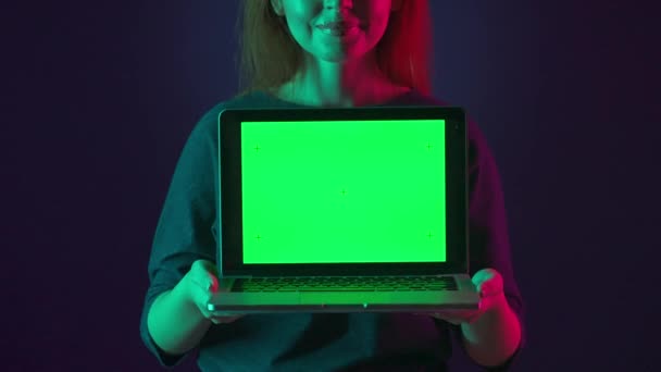 用粉色和绿色霓虹灯在蓝色背景上的女人手中拿着绿色屏幕的笔记本电脑 — 图库视频影像