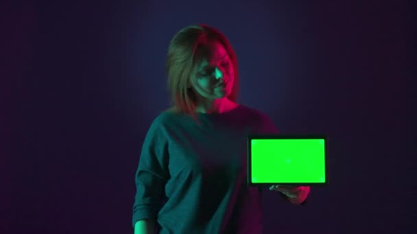 一个女人把食指指向平板电脑的绿色屏幕 然后竖起大拇指 一个女人的肖像 画室里有一个绿色屏幕的平板 蓝色背景上有粉色和绿色的霓虹灯 — 图库视频影像
