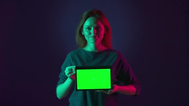 女性は緑色のタブレットの画面を閉じている ピンクと緑のネオンライトの青い背景にあるスタジオの緑色のスクリーンを使用した女性の肖像画 広告エリア — ストック動画