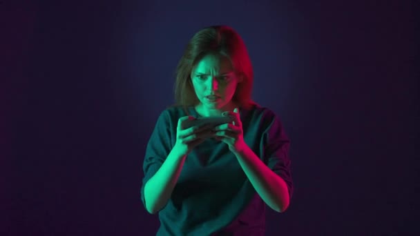 一个紧张的女人用她的智能手机玩网络游戏 赢得比赛并做出胜利的姿态 粉红和绿色霓虹灯下蓝色背景的工作室里的女人 — 图库视频影像