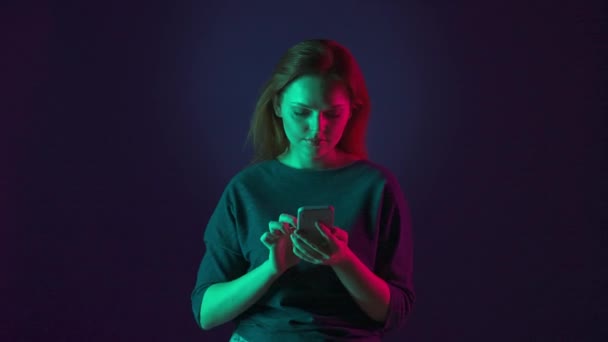 一个红头发的年轻女人在智能手机上输入一条信息 通过她在社交网络上的信息 与人交流 表现出一种好的姿态 在演播室里亮着粉色和绿色霓虹灯的女人 — 图库视频影像