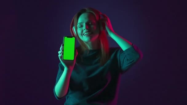 赤ワイヤレスヘッドフォンの女性は 緑色のスマートフォン画面を示しています 女性は目を閉じて音楽を聴き アプリを使用して 緑とピンクのネオンライトの青い背景にあるスタジオで — ストック動画