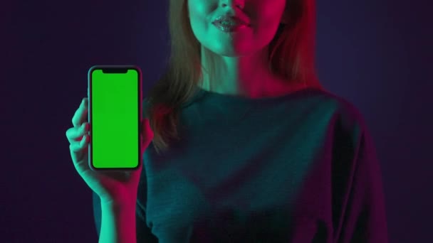ピンクと緑のネオンライトでスタジオで女性の緑色のスクリーンでスマートフォンが閉じます 女性は彼女のスマートフォンの緑色の画面で指を指差し 親指を示します — ストック動画