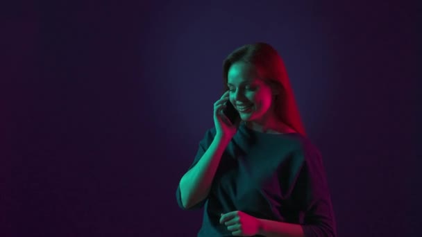 一个快乐的红头发女人在智能手机上说着情绪化的话 做着手势 在工作室里拿着智能手机 蓝色背景 粉色和绿色霓虹灯的女人 — 图库视频影像