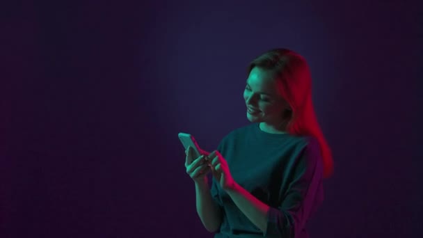 笑顔の若い赤毛の女性は スマートフォンでメッセージをタイプし ソーシャルネットワーク上で彼女のフィードをスクロールし 通信します ブルーのスタジオでピンクと緑のネオンライトで照らされた女性 — ストック動画
