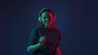 Beyaz kablosuz kulaklık takan genç, kızıl saçlı bir kadın DJ müziği dinliyor ve dans ediyor. Stüdyoda mavi arka planda pembe ve yeşil neon ışıklı bir kadın var.