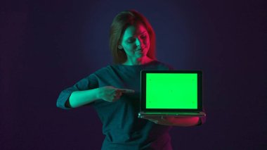 Bir kadın işaret parmağını yeşil bir dizüstü bilgisayara doğrultuyor. Stüdyoda yeşil ekranlı, pembe ve yeşil ışıklı mavi arka planda dizüstü bilgisayarı olan bir kadının portresi. Reklam platformu