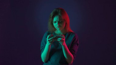Online bir oyunda akıllı telefonda kaybeden hayal kırıklığına uğramış bir kadının portresi. Stüdyoda mavi arka planda pembe ve yeşil neon ışıklı bir kadın var.